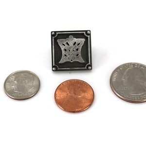 May include: A silver-colored metal pin with a square design featuring a stylized floral pattern. The pin is about the size of a US quarter.
