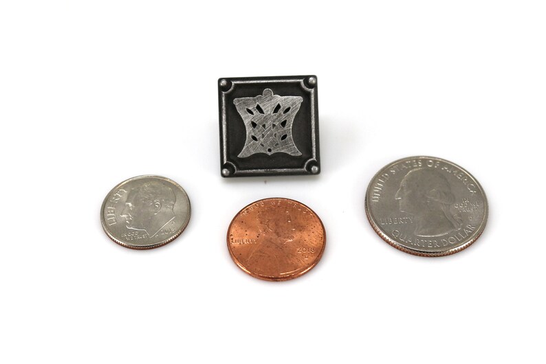 May include: A silver-colored metal pin with a square design featuring a stylized floral pattern. The pin is about the size of a US quarter.