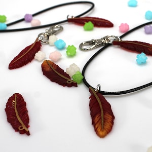 May include: Four red and gold feather charms on black cords. The charms are hanging from silver clasps. The charms are surrounded by small, colorful candies.