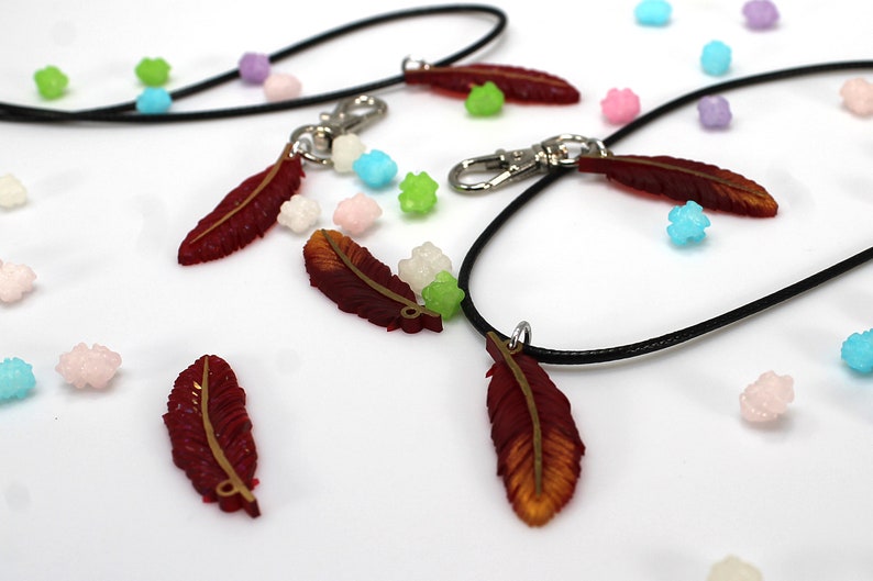 May include: Four red and gold feather charms on black cords. The charms are hanging from silver clasps. The charms are surrounded by small, colorful candies.