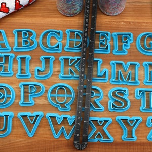May include: A set of 26 blue plastic alphabet cookie cutters on a wooden surface. The cutters are arranged in alphabetical order.