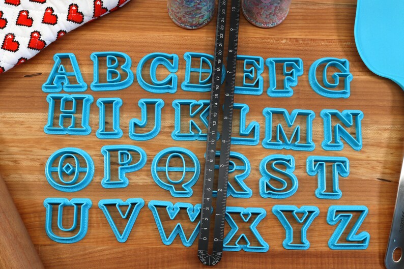 May include: A set of 26 blue plastic alphabet cookie cutters on a wooden surface. The cutters are arranged in alphabetical order.