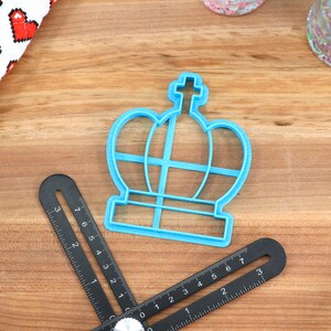 May include: A blue plastic crown-shaped cookie cutter on a wooden surface. The cutter has a cross on top and a base with a grid pattern.