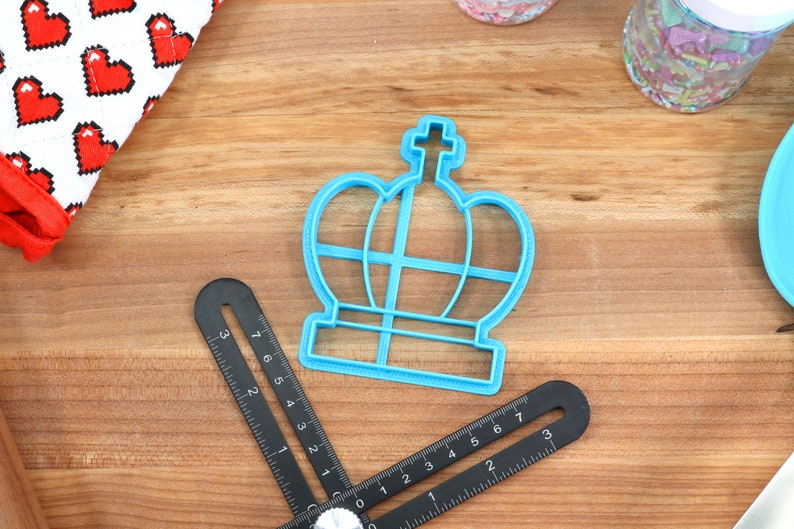 May include: A blue plastic crown-shaped cookie cutter on a wooden surface. The cutter has a cross on top and a base with a grid pattern.