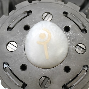 May include: A close-up of a metal gear with a white egg-shaped object in the center. The egg has a symbol on it that looks like a stylized wand with a crescent moon above it.