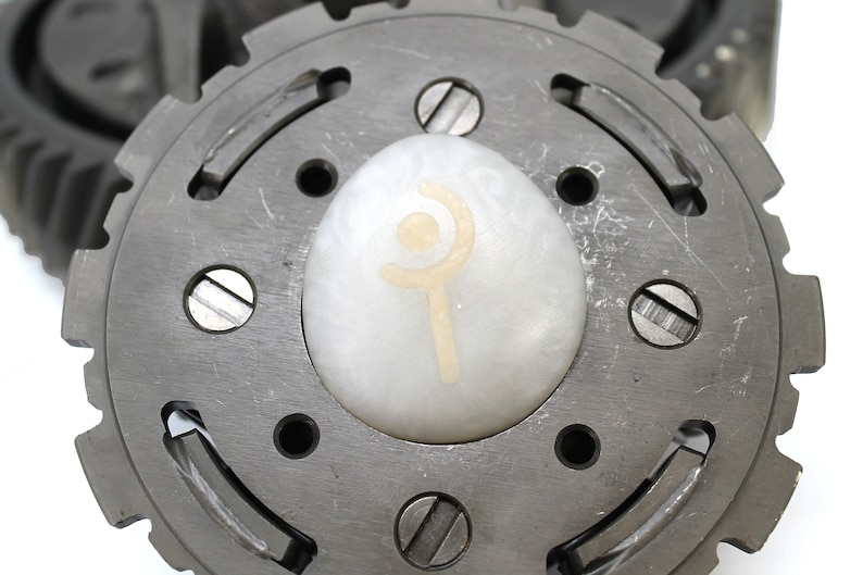 May include: A close-up of a metal gear with a white egg-shaped object in the center. The egg has a symbol on it that looks like a stylized wand with a crescent moon above it.