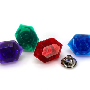 Puede incluir: Cuatro gemas de plástico en diferentes colores: morado, verde, rojo y azul. Las gemas tienen forma de octógono y un acabado brillante. Se incluye un alfiler plateado.