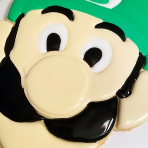 May include: A decorated cookie in the shape of a cartoon character's face. The cookie features a green hat with a white emblem, black mustache, and a cream-colored face. The eyes are white with black pupils, and the cookie is decorated with icing.