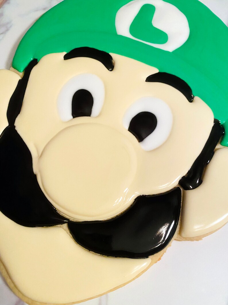 May include: A decorated cookie in the shape of a cartoon character's face. The cookie features a green hat with a white emblem, black mustache, and a cream-colored face. The eyes are white with black pupils, and the cookie is decorated with icing.