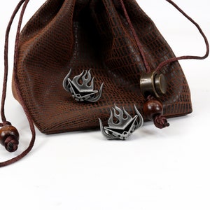 May include: Two silver metal dice with a flame design are sitting on a brown leather pouch with a drawstring. The pouch has a textured surface.
