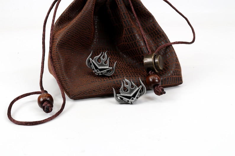 May include: Two silver metal dice with a flame design are sitting on a brown leather pouch with a drawstring. The pouch has a textured surface.
