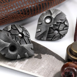 May include: Two small, silver-colored metal pendants with a textured, geometric design. Each pendant has two holes. The pendants are on a white surface.