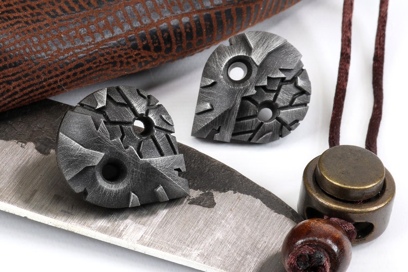 May include: Two small, silver-colored metal pendants with a textured, geometric design. Each pendant has two holes. The pendants are on a white surface.