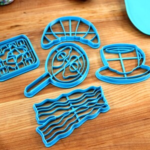 May include: A collection of bright blue plastic cookie cutters in various breakfast-themed shapes, including a waffle, fried egg, croissant, coffee cup, and bacon strip. The cutters are arranged on a wooden surface.