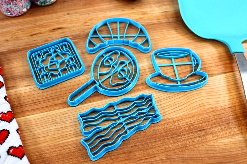 May include: A collection of bright blue plastic cookie cutters in various breakfast-themed shapes, including a waffle, fried egg, croissant, coffee cup, and bacon strip. The cutters are arranged on a wooden surface.