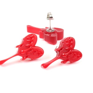 May include: A pair of red heart-shaped earrings with a dripping effect. The earrings are made of resin and have a silver post.