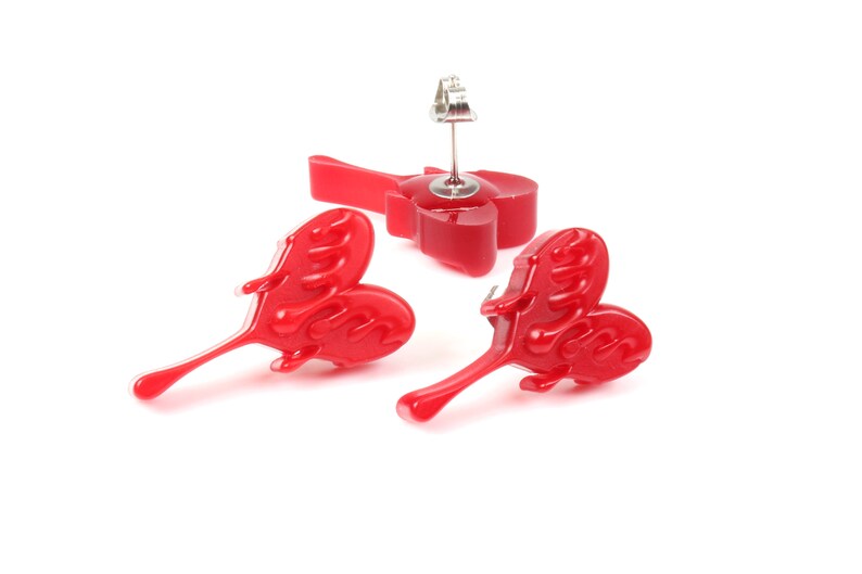 May include: A pair of red heart-shaped earrings with a dripping effect. The earrings are made of resin and have a silver post.