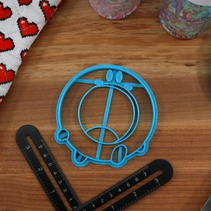 May include: A blue plastic cookie cutter shaped like a cartoon character with a circle and a line in the middle. The cookie cutter is on a wooden surface.