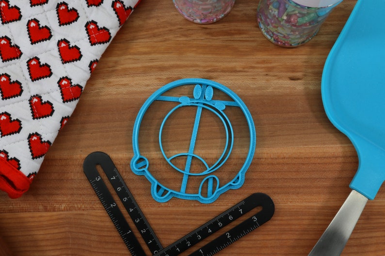 May include: A blue plastic cookie cutter shaped like a cartoon character with a circle and a line in the middle. The cookie cutter is on a wooden surface.