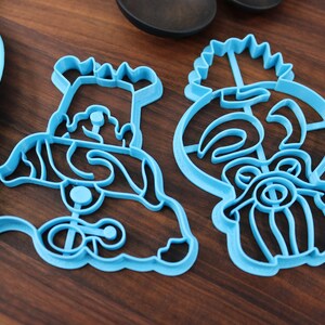 May include: Two blue plastic cookie cutters, one shaped like a cartoon character with a crown and the other shaped like a tiki head.