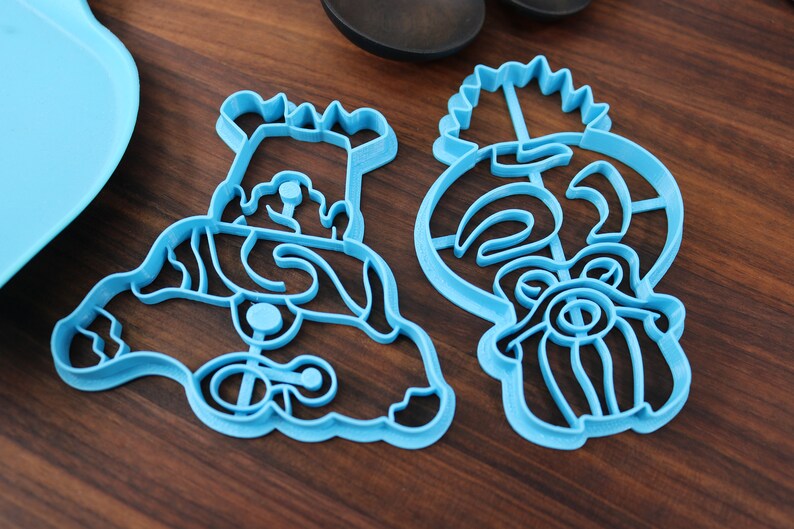 May include: Two blue plastic cookie cutters, one shaped like a cartoon character with a crown and the other shaped like a tiki head.
