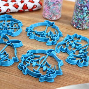 May include: Five blue plastic cookie cutters shaped like a cartoon character with a crown. The cutters are on a wooden surface.
