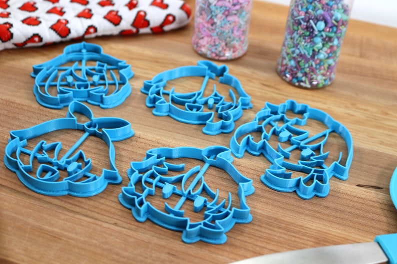 May include: Five blue plastic cookie cutters shaped like a cartoon character with a crown. The cutters are on a wooden surface.