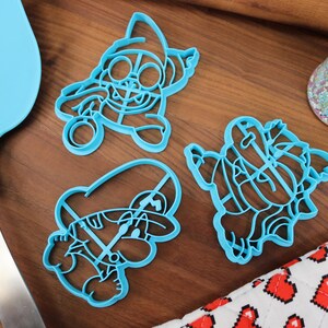 Op de afbeelding: Drie blauwe plastic koekjesvormen in de vorm van cartoonfiguren. De vormen liggen op een houten oppervlak.