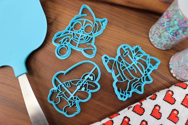 Op de afbeelding: Drie blauwe plastic koekjesvormen in de vorm van cartoonfiguren. De vormen liggen op een houten oppervlak.