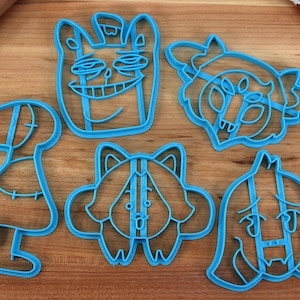 May include: Five blue plastic cookie cutters shaped like cartoon characters. The cutters are on a wooden surface.