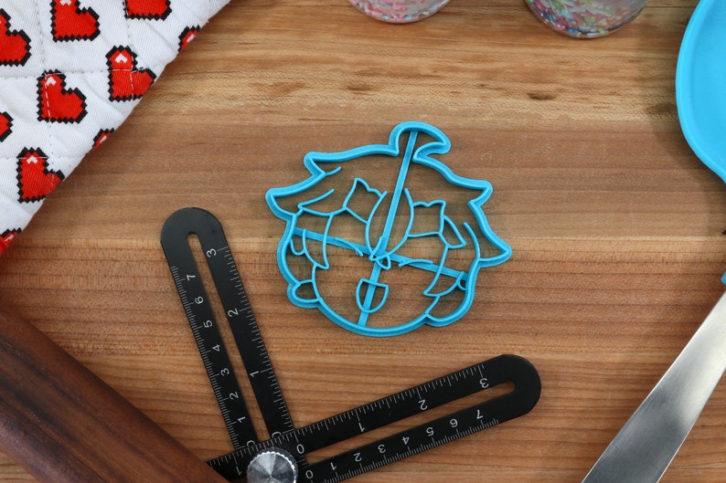 May include: A blue plastic cookie cutter shaped like a cartoon character with green hair and a shovel. The character has a surprised expression.