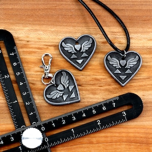 Puede incluir: Dos colgantes de plata en forma de coraz&oacute;n con un s&iacute;mbolo alado y un collar de cord&oacute;n negro. Un llavero de plata en forma de coraz&oacute;n con un s&iacute;mbolo alado y un anillo de plata.