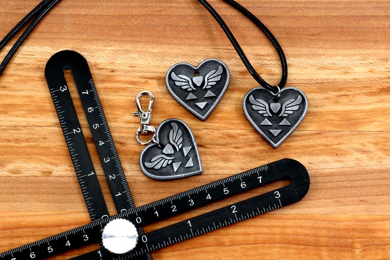 Puede incluir: Dos colgantes de plata en forma de coraz&oacute;n con un s&iacute;mbolo alado y un collar de cord&oacute;n negro. Un llavero de plata en forma de coraz&oacute;n con un s&iacute;mbolo alado y un anillo de plata.