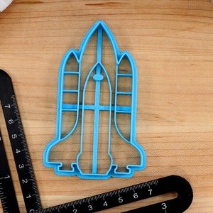 May include: A blue plastic cookie cutter shaped like a space shuttle. The cutter is on a wooden surface.