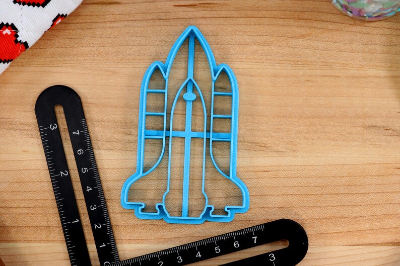 May include: A blue plastic cookie cutter shaped like a space shuttle. The cutter is on a wooden surface.
