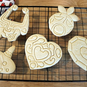 May include: Five intricately designed sugar cookies on a black wire cooling rack. The cookies are shaped like a heart, a video game item, a musical instrument, and two other video game items.