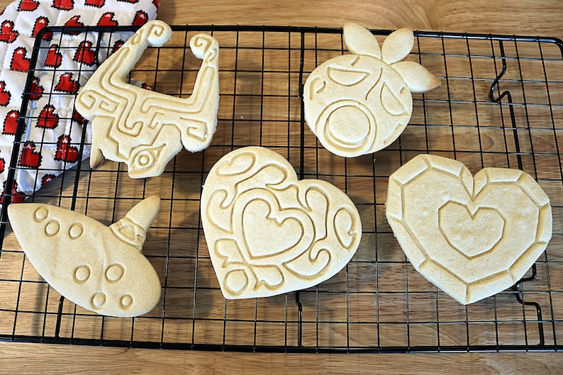 May include: Five intricately designed sugar cookies on a black wire cooling rack. The cookies are shaped like a heart, a video game item, a musical instrument, and two other video game items.