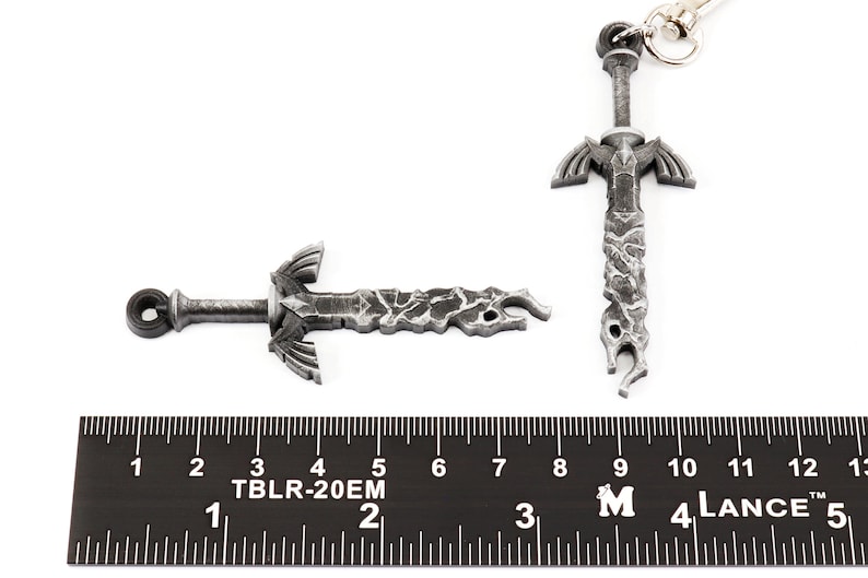 Może przedstawiać: Srebrny metalowy brelok do kluczy w kształcie miecza z fakturowaną powierzchnią. Miecz ma około 12,7 cm długości i ma pierścień na g&oacute;rze do przymocowania do breloka.
