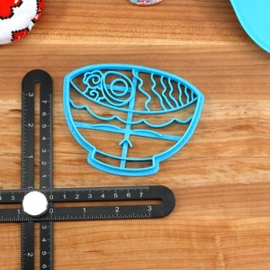 May include: A light blue plastic cookie cutter in the shape of a ramen bowl. The cutter features detailed designs of noodles, broth, and other ramen ingredients. The cutter is on a wooden surface with a ruler.