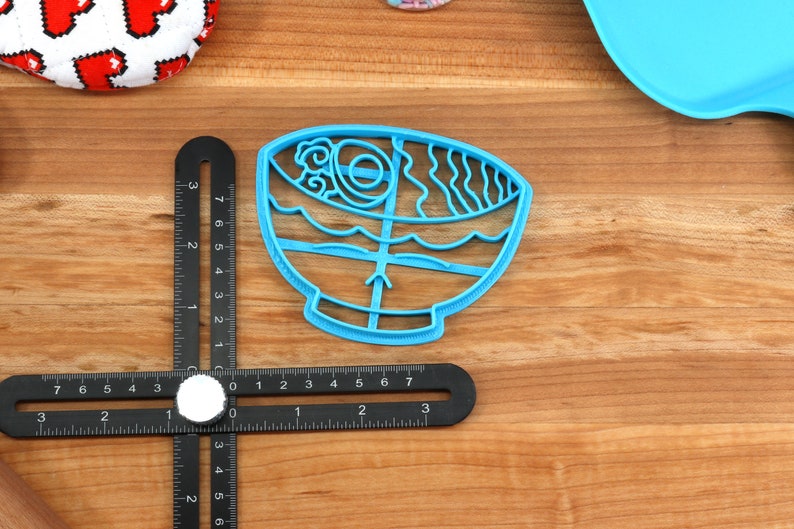 May include: A light blue plastic cookie cutter in the shape of a ramen bowl. The cutter features detailed designs of noodles, broth, and other ramen ingredients. The cutter is on a wooden surface with a ruler.