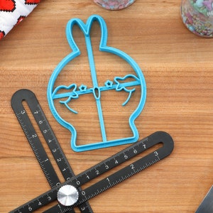 May include: A blue plastic cookie cutter shaped like a cartoon bunny rabbit with a round body and long ears. The cutter is on a wooden surface with a ruler and a rolling pin.