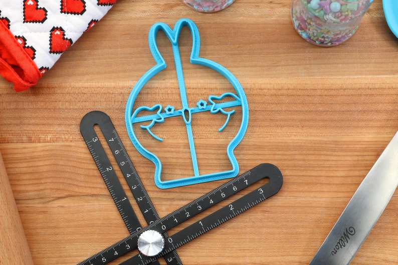 May include: A blue plastic cookie cutter shaped like a cartoon bunny rabbit with a round body and long ears. The cutter is on a wooden surface with a ruler and a rolling pin.