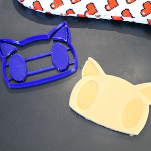 May include: A blue plastic cookie cutter shaped like a cat face, and a light brown cookie in the shape of a cat face.