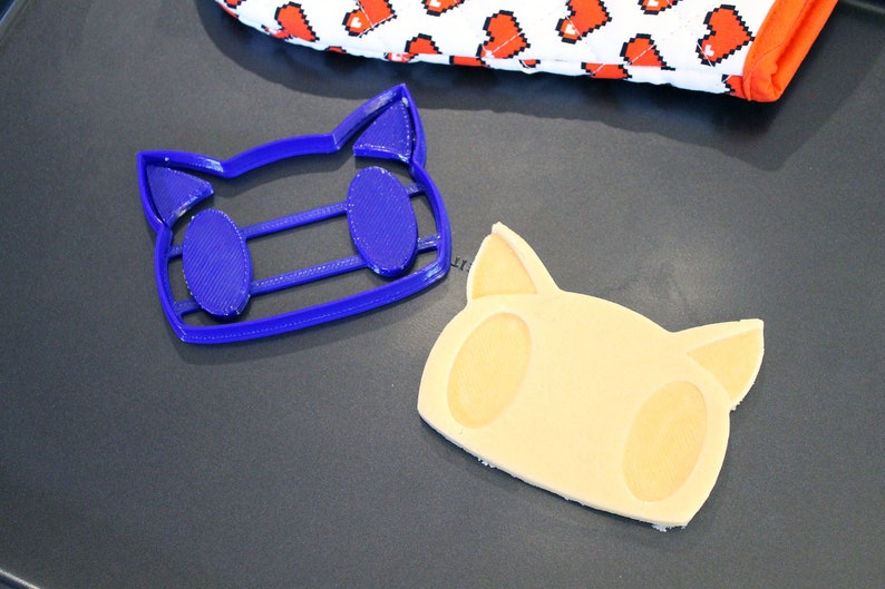 May include: A blue plastic cookie cutter shaped like a cat face, and a light brown cookie in the shape of a cat face.