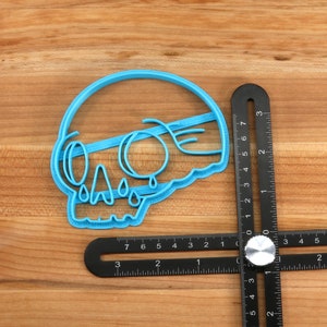 May include: A blue plastic cookie cutter shaped like a skull with dripping tears. The cutter is on a wooden surface next to a black ruler.