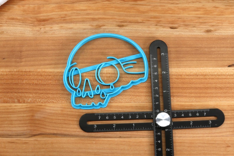 May include: A blue plastic cookie cutter shaped like a skull with dripping tears. The cutter is on a wooden surface next to a black ruler.