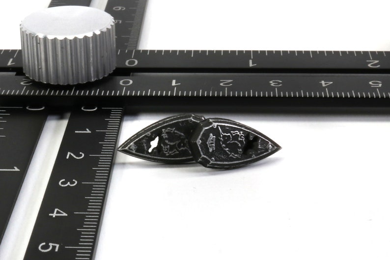 May include: Two antique black metal iron with intricate designs and a small hole on each side. The irons are sitting on a white surface with a black ruler in the background.