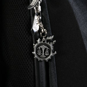 May include: A black backpack with a zipper pull featuring a silver charm with a stylized design. The charm has a circular shape with a tree and scales in the center.