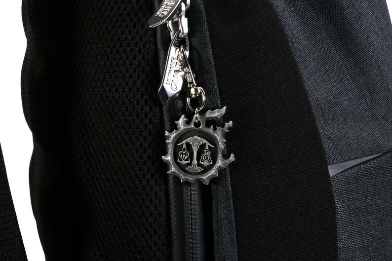 May include: A black backpack with a zipper pull featuring a silver charm with a stylized design. The charm has a circular shape with a tree and scales in the center.