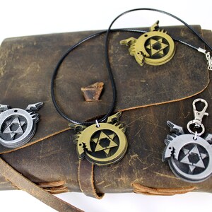 May include: Three metal pendants featuring a stylized winged serpent with a star of David inside. The pendants are in silver, gold, and bronze and are displayed on a brown leather journal.